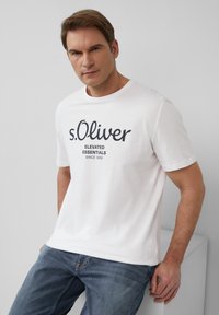 Biała bawełniana koszulka z czarnym nadrukiem "s.Oliver ELEVATED ESSENTIALS SINCE 1969" z przodu, w połączeniu z niebieskimi dżinsami.