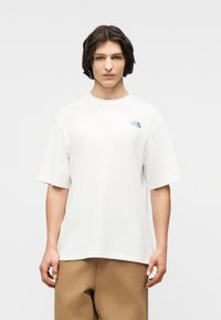 Νεαρός άνδρας με μεσαίου μήκους σκούρα μαλλιά, που φοράει λευκό t-shirt The North Face και καφέ παντελόνι, στέκεται μπροστά σε απλό ανοιχτόχρωμο φόντο.