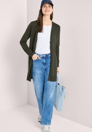 Donna che indossa un berretto blu navy, camicia bianca, cardigan lungo verde scuro, jeans blu, sneakers bianche, che tiene una borsa tote azzurro chiaro.