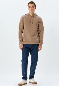 Sudadera beige con capucha y dobladillo acanalado, combinada con jeans de mezclilla azules y zapatillas blancas con suela color beige. Diseño sencillo, textura suave.
