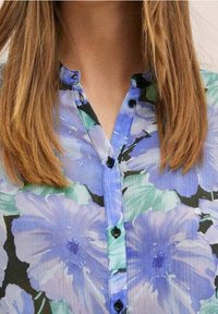 Blouse à fleurs avec de grands motifs floraux bleus et verts, fermeture à boutons noirs à l'avant, et une texture de tissu lisse.