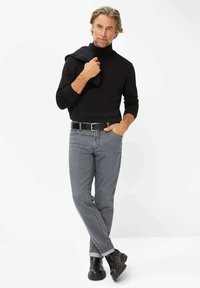 Schwarzer Rollkragenpullover, graue Jeans, schwarzer Gürtel und schwarze Stiefeletten. Mann, der eine dunkelgraue Jacke lässig über der Schulter hält.