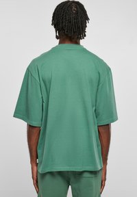 Tricou oversized verde din bumbac; mâneci scurte, guler rotund, textură fină. Detaliu de broderie notabil la tiv și eticheta laterală.