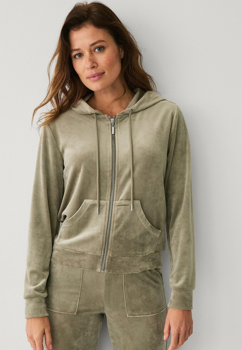 Haut à capuche zippé vert olive en tissu doux et velouté. Il présente une capuche, des poches avant et des cordons de serrage avec des accessoires en argent. Texture lisse.