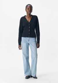 Kvinna står klädd i en svart knappad kofta, ljusblå vida jeans och svarta skor mot en helt vit bakgrund.