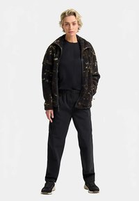 Schwarz-goldene, flauschige Stehkragenjacke mit Reißverschluss und zwei Seitentaschen, kombiniert mit schwarzen, schmal zulaufenden Hosen und dunklem Schuhwerk.