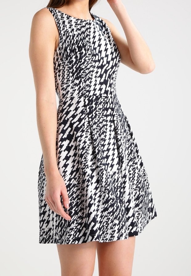 Mustat ja valkoiset houndstooth-kuvioiset ilmanhihoja oleva mekko, jossa on hyvin istuva yläosa ja liehuva helma, pukeutuneena seisovalle henkilölle.