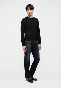 Mies seisoo valkoisella taustalla yllään musta pitkähihaisen neulepaita, tummansiniset farkut ja mustat pukukengät.