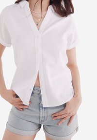 Chemise blanche à manches courtes avec boutons, en tissu léger, associée à un short en denim clair. Caractérisée par un col classique et des manches retroussées.