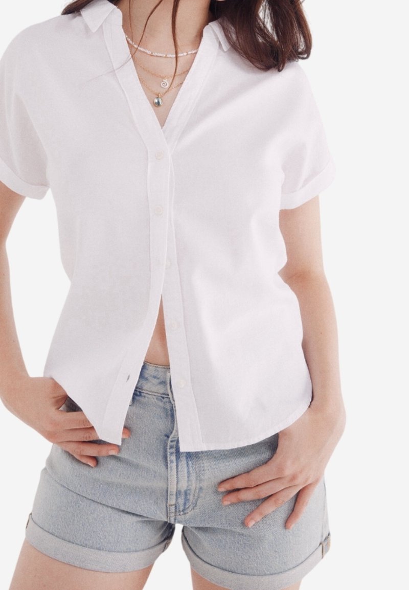 Chemise blanche à manches courtes avec boutons, en tissu léger, associée à un short en denim clair. Caractérisée par un col classique et des manches retroussées.