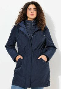 Giacca lunga blu navy con cappuccio, vita elastica, tasche con zip e chiusura a bottoni a pressione; presenta un rivestimento interno trapuntato.