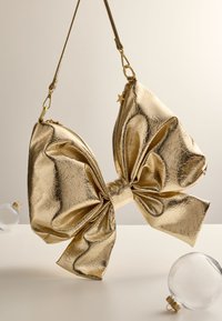 Kultainen metallinhohtoinen rusettimuotoinen käsilaukku, jossa kiiltävä pinta, suuri rusetti edessä ja vetoketjullinen osasto. Olkahihna on kiinnitetty kullanvärisellä metallilla.