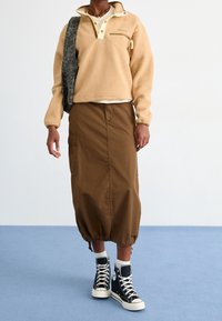 Beżowy polarowy sweter z zapięciem na napy i kieszenią na piersi, zestawiony z brązową spódnicą midi z bocznymi kieszeniami i ściągaczem na dole, noszony z czarnymi trampkami za kostkę.
