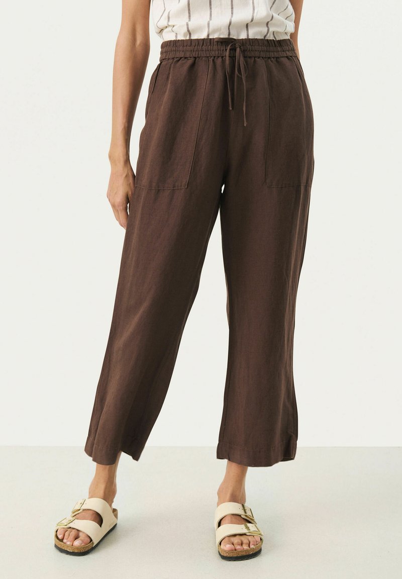 Pantalons en lin marron au look ample, avec une ceinture élastique et deux poches avant. Associés à des sandales beiges avec un design à boucle.
