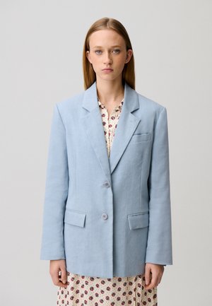 Giovane donna con capelli biondi lisci che indossa un blazer azzurro su misura sopra un vestito a fiori su uno sfondo semplice.