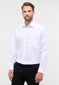 Camicia bianca a maniche lunghe con bottoni, realizzata in tessuto morbido, caratterizzata da una tasca sul petto e bottoni neri lungo la parte anteriore e i polsini.