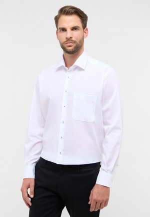 Witte, langslavige blouse gemaakt van gladde stof, met een enkele borstzak en zwarte knopen langs de voorkant en de manchetten.