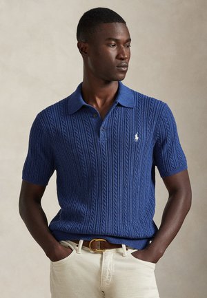 Marineblaues Polohemd mit kurzen Ärmeln, Kabelstrickmuster, kontrastierendem Kragen und gesticktem Logo. Kombiniert mit hellen Hosen.