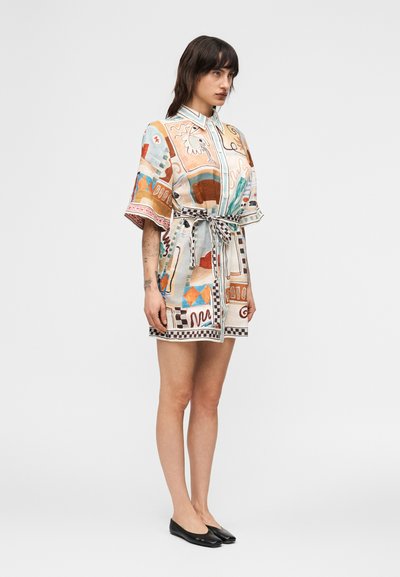 Νεαρή γυναίκα που στέκεται πλαγίως, φορώντας ένα φόρεμα με ζώνη, κοντά μανίκια και σχέδιο, μαζί με μαύρα φλατ παπούτσια, σε απλό φόντο.