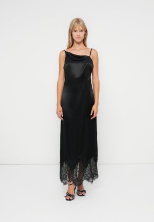 Abito maxi in satin nero con scollatura asimmetrica, dettaglio in pizzo all'orlo e spalline regolabili. Abbinato a sandali neri a pois.