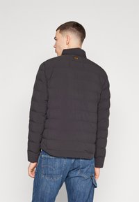 Jacheta puffer neagră are umplutură matlasată, guler înalt și un mic eticheta logo pe spate, fiind asortată cu blugi albaștri.