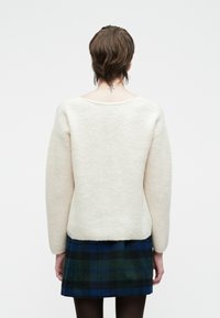 Kremfarget strikkegenser med lange ermer, rund hals, og myk tekstur, kombinert med et mørkegrønt og blått rutete skjørt.