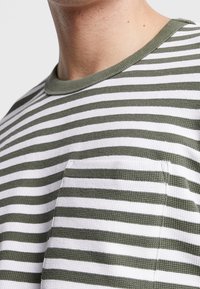 Gestreiftes T-Shirt mit horizontalen olivgrünen und weißen Linien, Rundhalsausschnitt, strukturiertes Material, mit einer aufgesetzten Tasche auf der linken Seite.