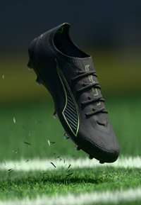 Crampons de football noirs avec une texture nervurée, des accents verts et une forme épurée. Comprend six œillets et un logo surélevé sur le côté.