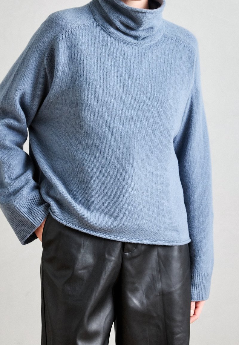 Personne portant un pull à col roulé bleu clair à manches longues et un pantalon en cuir noir avec une main dans la poche.