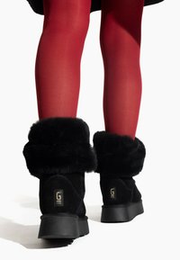 Botas de tobillo de ante negro con un suave collar de pelo sintético negro, suela de goma plana y detalle del logo en el talón. Se ven medias rojas.