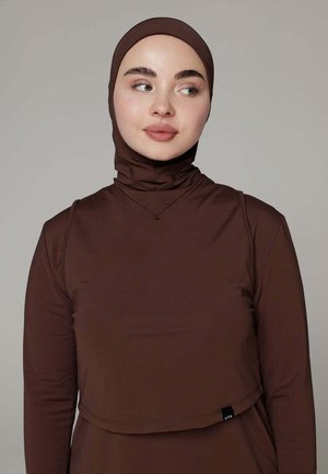 Jonge vrouw die een aansluitende bruine hijab en bijpassende longsleeve draagt, licht naar links kijkend tegen een effen achtergrond.