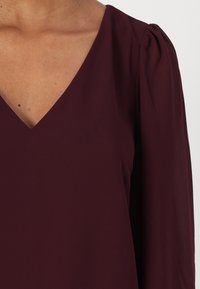 Bordo bluza s V-izrezom, dugim napuhanim rukavima i glatkom teksturom. Tkanina izgleda lagano i lijepo se drapira.