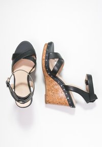 Czarne skórzane sandały na koturnie z korkowym obcasem. Krzyżujące się paski i regulowany pasek na kostkę. Wyraźne srebrne ćwieki wzdłuż krawędzi obcasa.