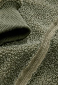 Struktureret olivengrøn fleece i et blødt og varmt udseende med en glat kontrastmanchet, der viser en synlig søm langs kanten.