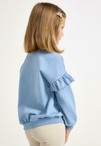 Jasnoniebieska bluza z falbankami na rękawach, zaokrąglonym dołem i ściągaczami. Modelka odwrócona tyłem; widoczne beżowe spodnie o teksturowanej fakturze.