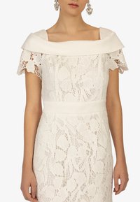 Abito bianco in pizzo con scollatura strutturata a spalle scoperte, motivi floreali e silhouette aderente, accentuato da una cintura abbinata.