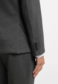 Gray tailored suit with a textured finish. The jacket features a buttoned cuff and fits closely at the waist. White shirt cuffs visible beneath.