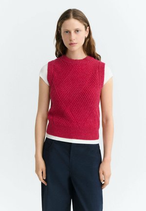 Jonge vrouw met een rood gebreide mouwloze trui over een wit t-shirt en donkere broek, staand tegen een effen lichte achtergrond.