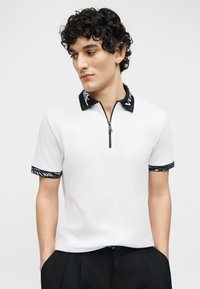 Νεαρός άνδρας με σγουρά σκούρα μαλλιά που φοράει λευκό κοντομάνικο πουκάμισο με μαύρο γιακά και τελειώματα στα μανίκια, κοιτάζοντας ελαφρώς προς τα αριστερά.