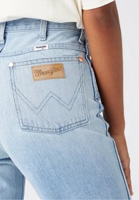 Jeans de mezclilla azul claro con parche de cuero Wrangler y etiqueta blanca, usados por una persona en camisa blanca, mostrando el detalle del bolsillo trasero y la costura lateral.