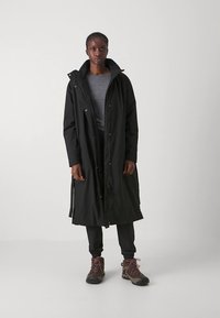 Svart lång kappa med huva, tillverkad av vattentätt tyg. Har en dragkedja framtill, dragsko i midjan och sidofickor. Bärs över en grå skjorta.