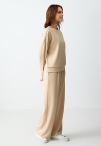 Beige sweatshirt en wijde broek van zacht, gewatteerd materiaal. De outfit heeft een ontspannen pasvorm en contrasterende afwerking op de schouders.