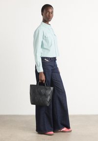 Svart vävd läderhandväska med en strukturerad form. Modellen bär en ljusblå randig skjorta och vida mörk denimjeans med rosa sneakers.