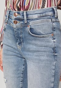 Mujer con jeans de mezclilla de tiro alto en azul claro, con dos botones de cobre, y una camisa a rayas multicolor metida por dentro.