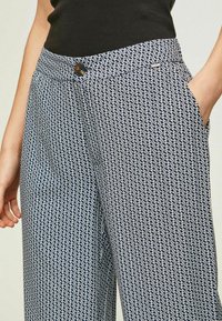 Frau trägt marineblauen und weißen gemusterten Hose mit einem knöpfbaren Bund, schwarzem Oberteil und hat die Hand in der Hosentasche.