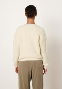 Kremo spalvos megztinis su struktūrizuotu, ribuotu raštu ir nuleistais pečiais, derinamas su aukšto liemens, žemės tonų kelnėmis.