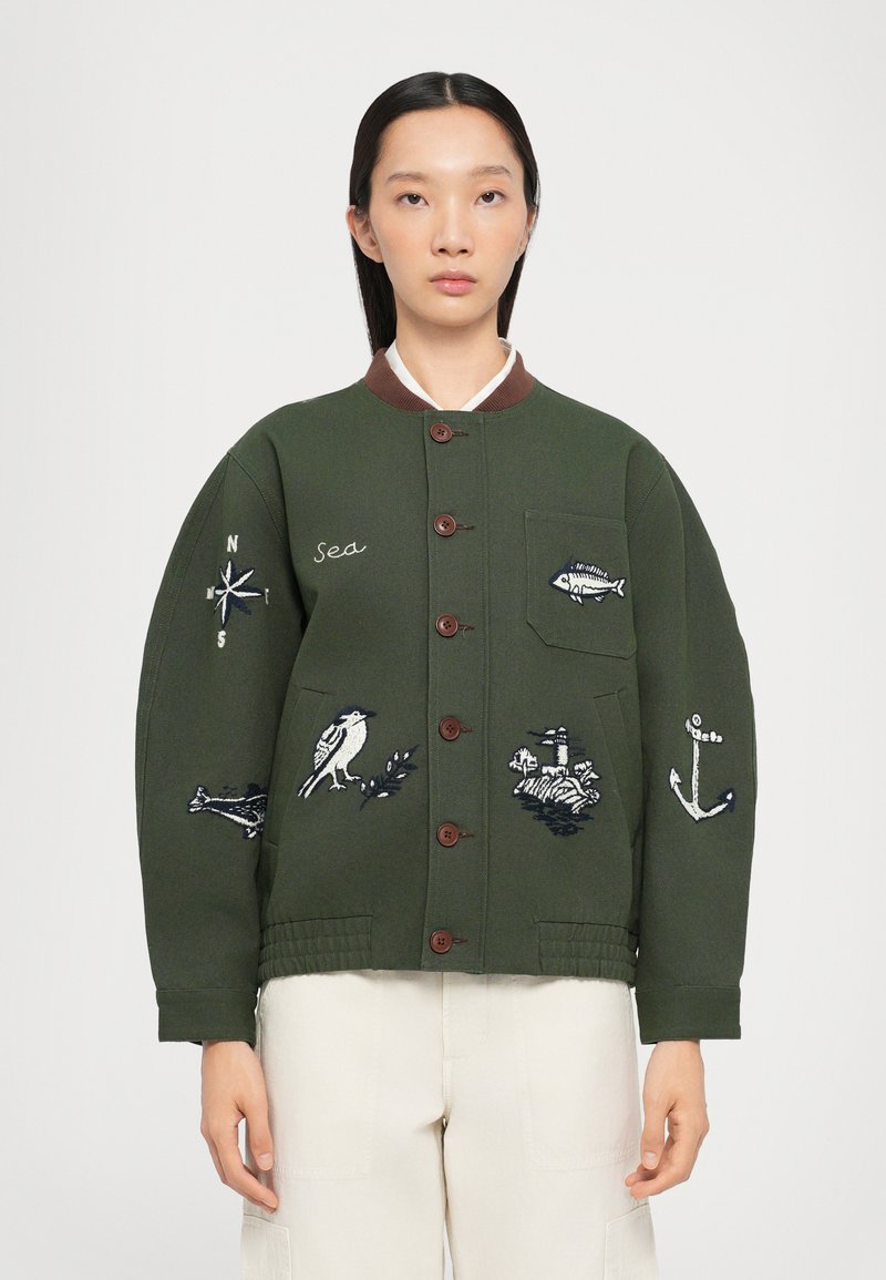 Chaqueta de algodón verde con cuello marrón, con motivos marinos bordados, incluyendo un pájaro, un pez, un ancla y una brújula. Botones en el frente y un bolsillo.