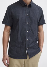 Camisa de manga corta de un azul oscuro, hecha de tejido texturizado, con cuello abotonado y botones de madera a lo largo del cierre frontal.