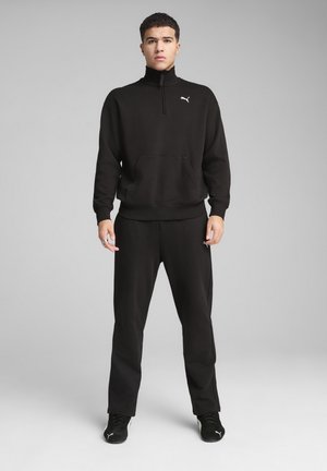 Schwarzer Reißverschluss-Sweatshirt mit hohem Kragen, Kängurutasche und Puma-Logo. Kombiniert mit passenden schwarzen Jogginghosen und schwarzen Sneakern.