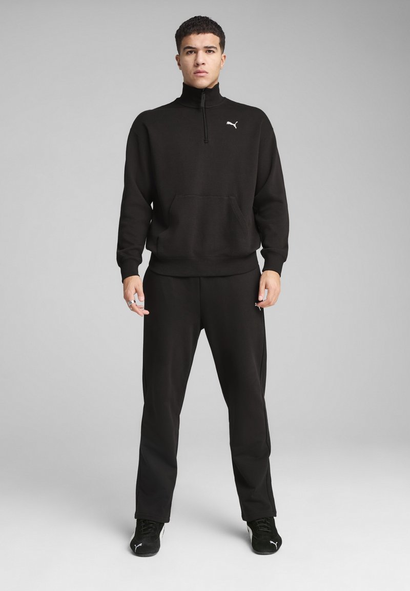 Sort hættetrøje med lynlås og høj krave, kængurulomme og Puma-logo. Sammen med matchende sorte sweatpants og sorte sneakers.